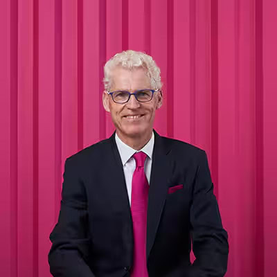A man with curly white hair, glasses, and a dark suit with a pink tie sits in front of a vertical pink striped background, smiling at the camera.