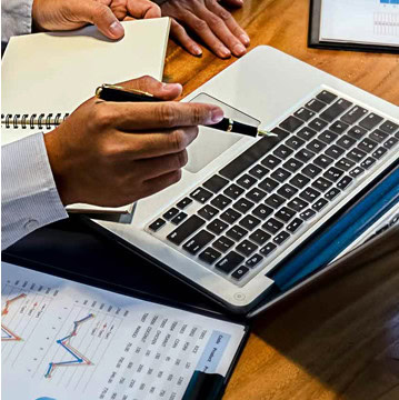 Person pointing at a laptop screen with a pen. Nearby, a notebook, business charts, and another person's hands on a wooden table.