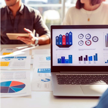 Laptop displaying graphs and charts on a table with printed reports; two people discussing in the background.
