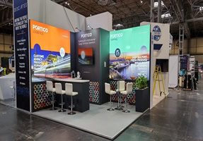 Trade show booth with colorful display panels, white chairs, a standing counter, and promotional material on tables.