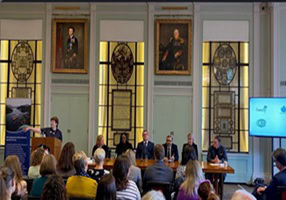 A panel of six people seated at a table in a decorated room. An audience watches, and a speaker stands at a podium. A screen displays logos.