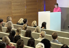 A person speaks at a white podium to an audience seated in rows, with wooden paneling and a blue light in the background.