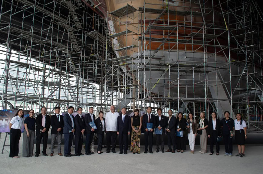 A group of people in business attire stands in front of a large structure covered in scaffolding, posing for a group photo at a construction site.