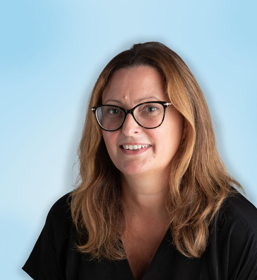 A woman with long brown hair and glasses, wearing a black top, smiles in front of a light blue background.