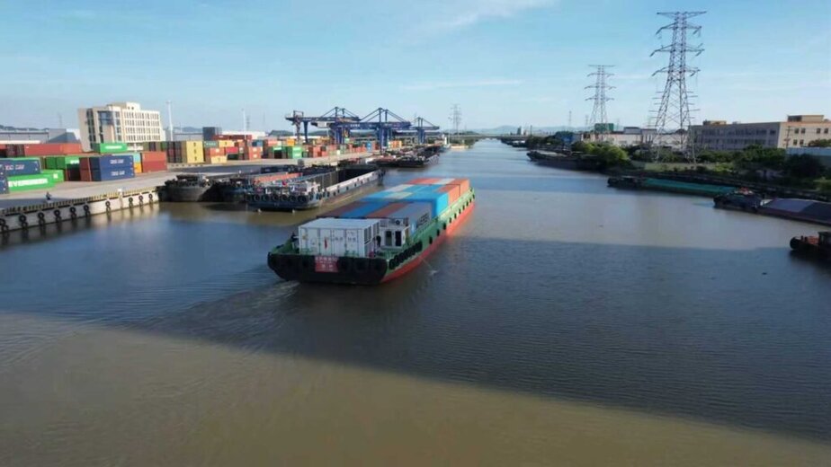 A cargo ship carrying containers moves down a wide river flanked by industrial buildings, cranes, and power lines under a clear blue sky.