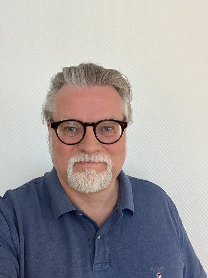 A man with grey hair, a beard, and glasses is wearing a blue polo shirt and smiling at the camera in front of a light patterned wall.