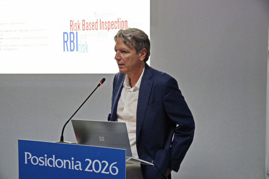 A man in a suit speaks at a lectern labelled “Posidonia 2026”, with a presentation slide about Risk-Based Inspection displayed behind him.