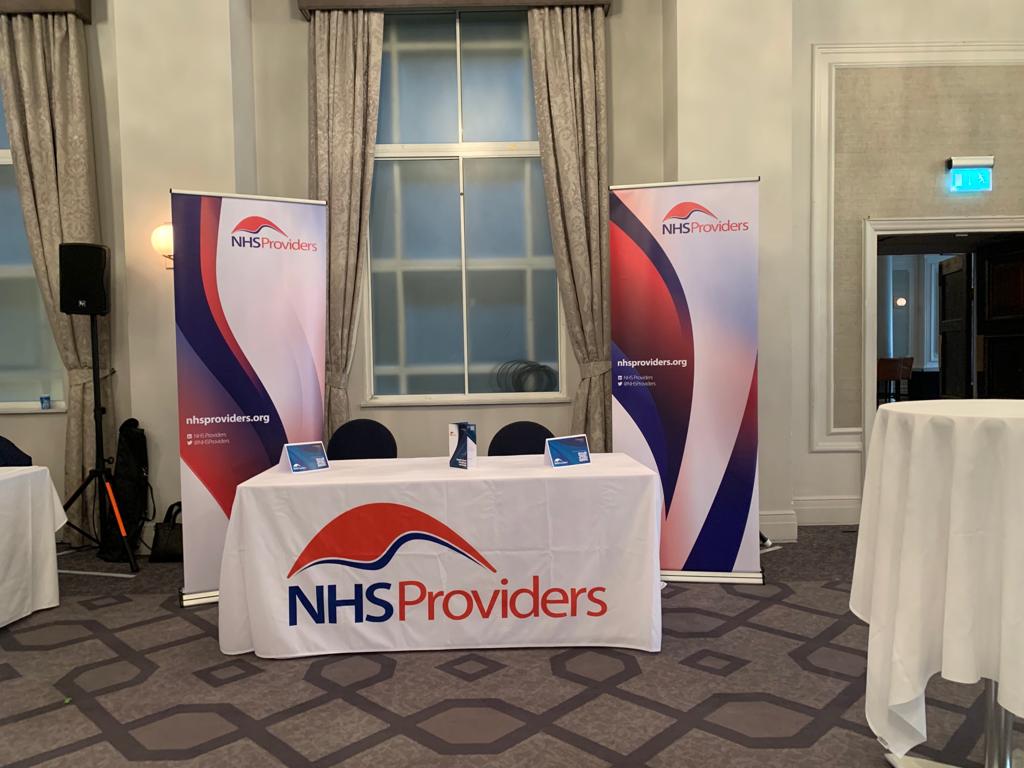 A conference setup with a table covered by an NHS Providers cloth, flanked by two banners. The table holds three brochures.