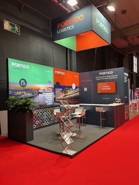 Trade show booth with Portico Logistics branding. Includes brochures, a counter with high chairs, display screens, and informational panels on shipping and cargo logistics.