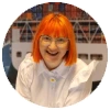 A person with orange hair and glasses, wearing a white shirt, smiles in front of a blurry background featuring a ship.